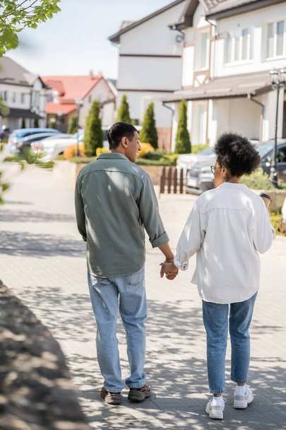 homme afro-américain souriant tenant la main de sa petite amie avec café à emporter tout en marchant à l'extérieur - Photo, image