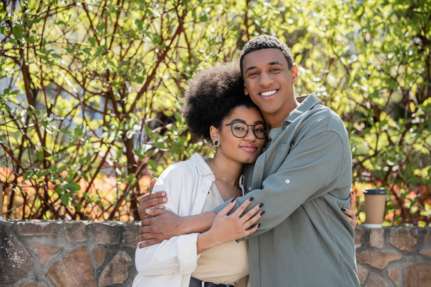 smiling african american man hugging girlfriend and looking at camera near coffee to go outdoors - Photo, Image