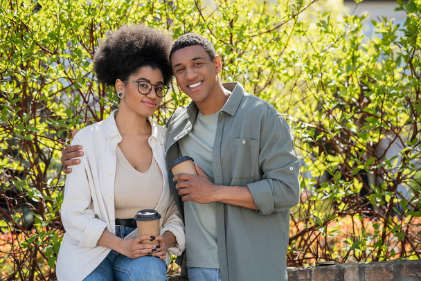 χαμογελαστό ζευγάρι Αφροαμερικανών που κρατούν καφέ και αγκαλιάζουν την ύπαιθρο το καλοκαίρι - Φωτογραφία, εικόνα