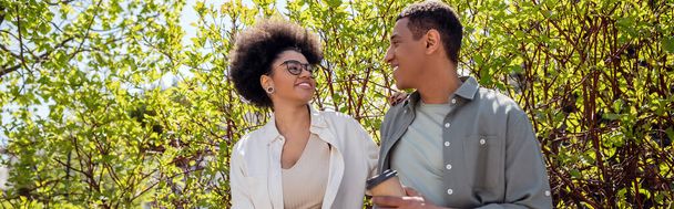 sonriente hombre afroamericano con café para ir a hablar con su novia cerca de los árboles al aire libre, pancarta - Foto, Imagen