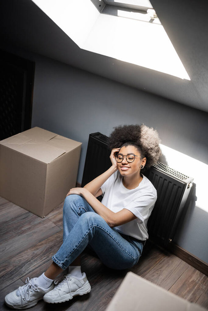 vista ad alto angolo di donna afro-americana sorridente seduta vicino a scatole di cartone in soffitta in casa hew - Foto, immagini