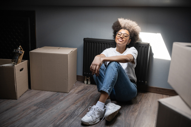 szczęśliwy Afroamerykanka kobieta siedzi w słońcu w pobliżu kartonowe pudełka na poddaszu w nowym domu - Zdjęcie, obraz