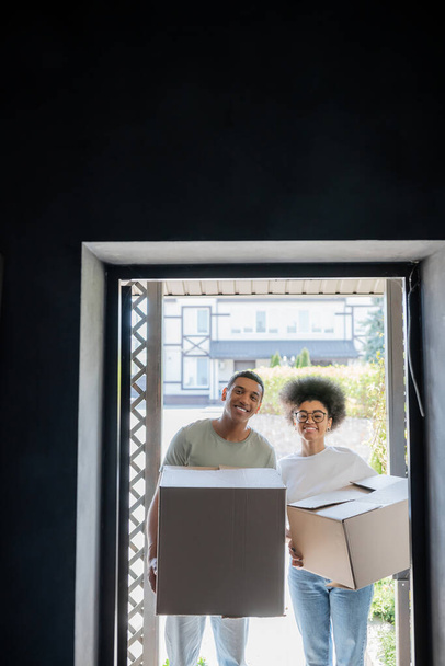 sonriente afroamericano pareja sosteniendo cajas de cartón y mirando a la cámara cerca de puerta en nueva casa - Foto, imagen