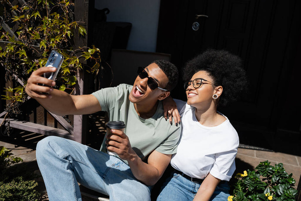 cheerful african american couple with coffee taking selfie on smartphone near new house - Foto, imagen