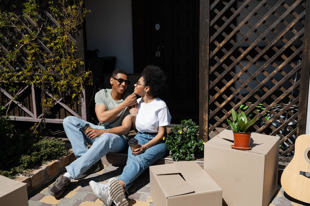 θετική αφρικανική αμερικανικό ζευγάρι με καφέ για να πάει κάθεται κοντά σε χαρτοκιβώτια και το νέο σπίτι - Φωτογραφία, εικόνα