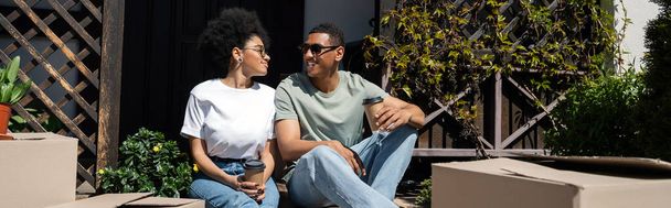 幸せなアフリカ系アメリカ人男性は,カートンボックスや新しい家,バナーの近くでコーヒーとガールフレンドと話す - 写真・画像