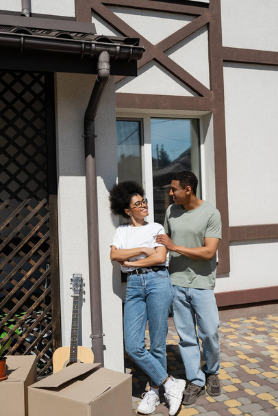 улыбающаяся африканская американская пара, стоящая рядом с картонными коробками и новым домом на открытом воздухе - Фото, изображение