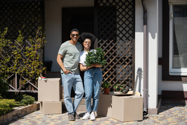 positif couple afro-américain avec plante d'intérieur debout près de boîtes en carton et nouvelle maison - Photo, image