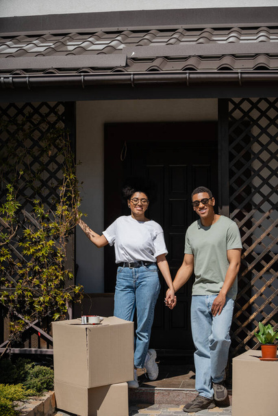 ευτυχισμένο ζευγάρι της Αφρικής κρατώντας το χέρι κοντά σε χαρτοκιβώτια και στη βεράντα του νέου σπιτιού - Φωτογραφία, εικόνα