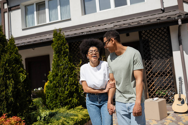 excited african american woman standing near boyfriend and new house on background outdoors - Photo, Image