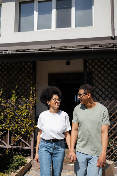 smiling african american man in sunglasses holding hand of girlfriend near new house outdoors - Foto, Imagem