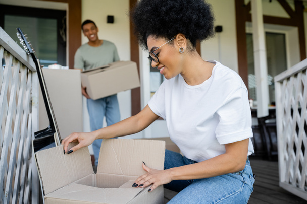 cheerful african american woman unpacking carton box near blurred boyfriend and new house - Photo, Image