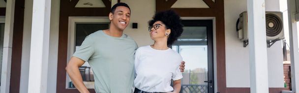pareja afroamericana positiva abrazándose mientras está de pie en el porche cerca de nueva casa, bienes raíces, pancarta - Foto, Imagen