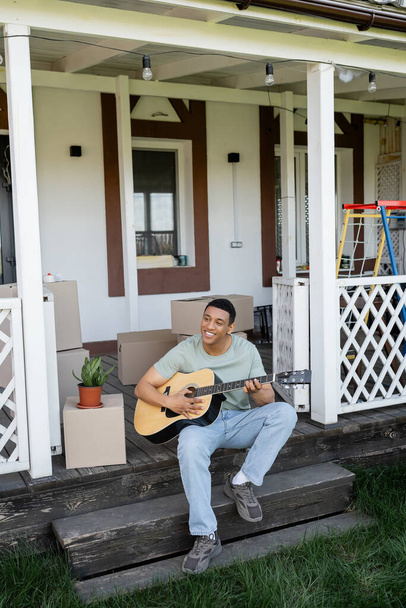 fröhlicher afrikanisch-amerikanischer Mann spielt Akustikgitarre auf Veranda in der Nähe von Boxen und neuem Haus - Foto, Bild