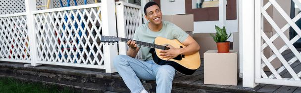 hombre afroamericano feliz tocando la guitarra acústica cerca de cajas de cartón en el porche de la nueva casa, bienes raíces, bandera - Foto, imagen