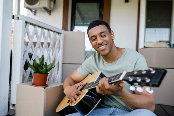 新しい家のポーチのパッケージの近くでアコースティックギターを演奏する楽しいアフリカ系アメリカ人男性 - 写真・画像
