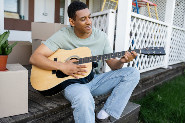 Веселый африканский американец играет на акустической гитаре рядом с картонными коробками и новым домом - Фото, изображение