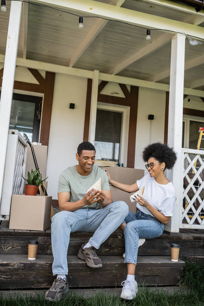 positive african american couple with sandwiches and coffee talking on porch of new house - Photo, Image