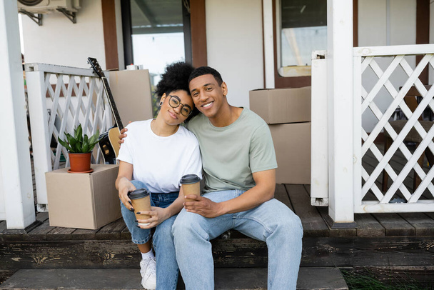 lächelnd afrikanisch-amerikanisches Paar mit Kaffee zum Umarmen, während es auf der Couch des neuen Hauses sitzt - Foto, Bild