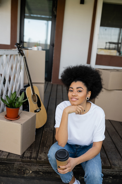 pozytywna Afroamerykanka trzyma kawę na wynos i patrzy w kamerę w pobliżu nowego domu - Zdjęcie, obraz