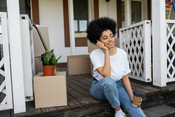 pleased african american woman holding coffee to go while sitting near carton boxes and new house - Photo, Image