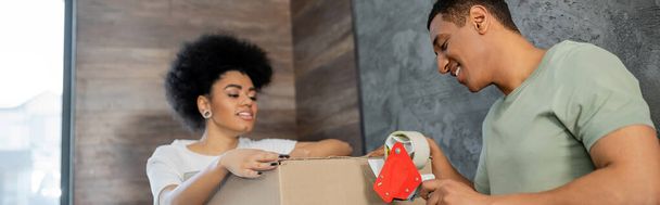 улыбающийся африканский американец держит клейкую ленту рядом с коробкой и подругой во время переезда, баннер - Фото, изображение