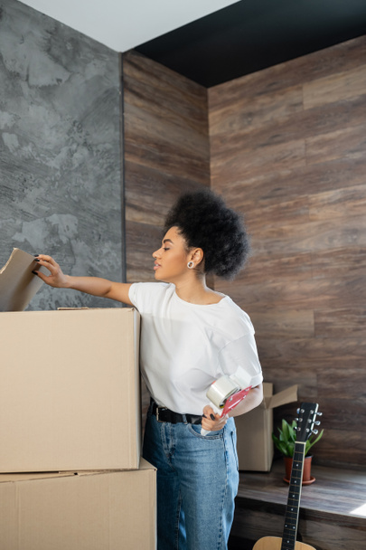side view of african american woman holding adhesive tape near carton boxes during moving - Photo, Image