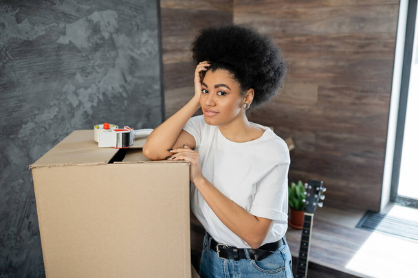 glimlachende Afro-Amerikaanse vrouw op zoek naar camera in de buurt van kartonnen doos in de woonkamer in nieuw huis - Foto, afbeelding