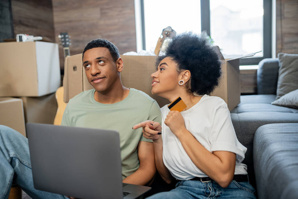 afro-américaine détenant une carte de crédit et pointant vers un ordinateur portable près de petit ami lors de la relocalisation - Photo, image