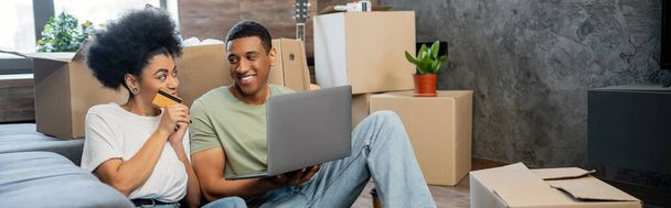 positief Afrikaans Amerikaans paar met laptop en creditcard tijdens verhuizing in nieuw huis, banner - Foto, afbeelding