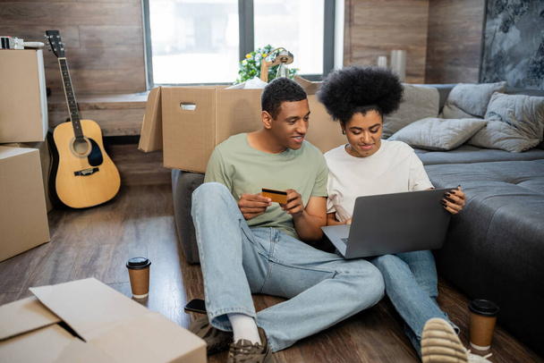 african american man holding credit card near girlfriend with laptop and boxes in new house - Fotó, kép