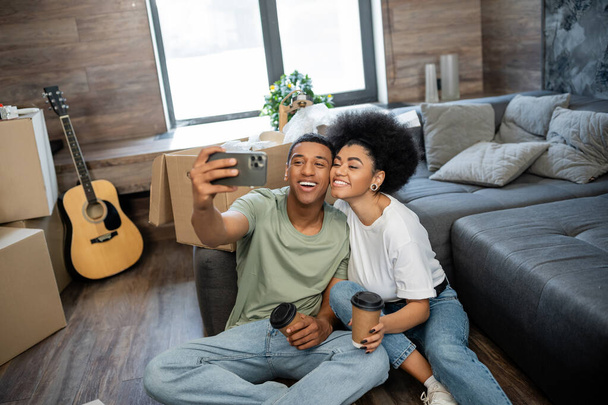 gelukkig Afrikaans Amerikaans paar nemen selfie en het houden van koffie in de buurt van pakketten in nieuwe woonkamer - Foto, afbeelding