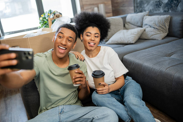 アフリカ系アメリカ人のカップルは,スマートフォンでセルフィーを撮り,新しい家でコーヒーを保持しながら笑っています - 写真・画像
