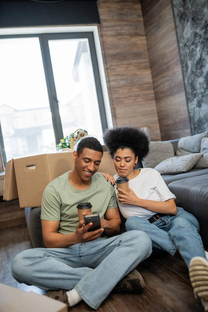 θετικοί αφρικανοί ζευγάρι με καφέ χρησιμοποιώντας smartphone κοντά σε πακέτα σε νέο σπίτι - Φωτογραφία, εικόνα