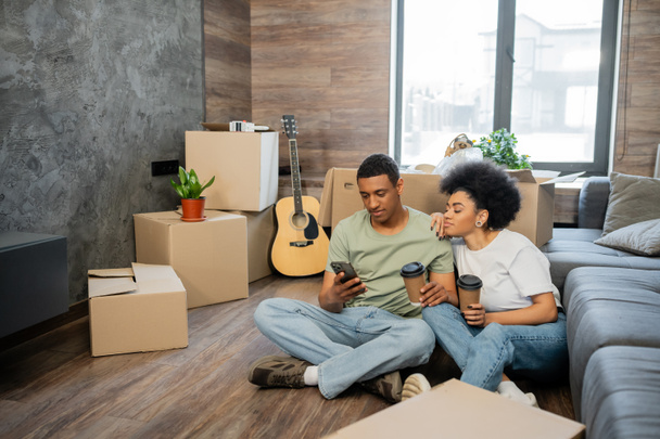 african american couple using cellphone and holding coffee near packages in new living room - Photo, Image