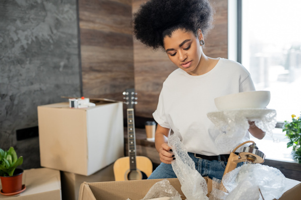 mulher afro-americana desempacotando utensílios de mesa perto de caixa de papelão em nova casa - Foto, Imagem