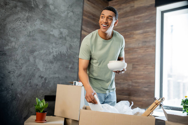 hombre afroamericano positivo sosteniendo vajilla mientras desempaqueta cajas de cartón en casa nueva - Foto, imagen