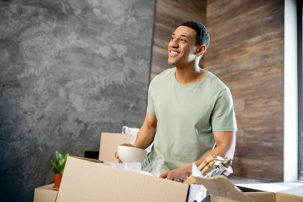 alegre afroamericano hombre mirando hacia otro lado mientras desembalaje paquetes en nueva casa - Foto, imagen
