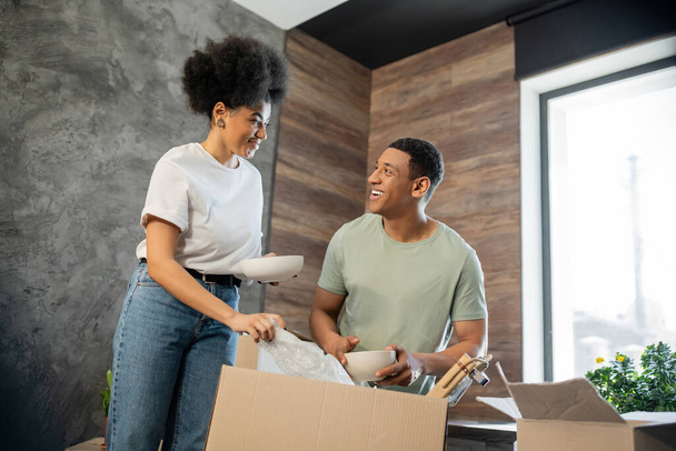 sonriente afroamericana mujer sosteniendo placa y hablando con novio desembalaje caja en nueva casa - Foto, Imagen
