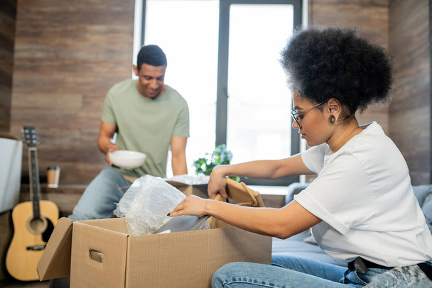 mujer afroamericana desembalaje caja de cartón cerca borrosa novio durante la reubicación en nueva casa - Foto, Imagen