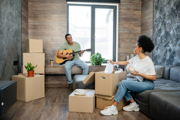 lächelnder afrikanisch-amerikanischer Mann spielt Akustikgitarre in der Nähe von Freundin und Kartons in neuem Haus - Foto, Bild