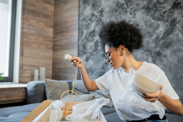vista lateral de la mujer afroamericana sosteniendo la placa y el alambre mientras desempaca la caja en una casa nueva - Foto, imagen