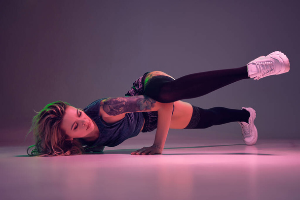 Young beautiful caucasian blonde girl with tattoo on her body, wearing velour blue booty shorts and black stockings dancing twerk lying down with raised leg, pink background - Photo, Image