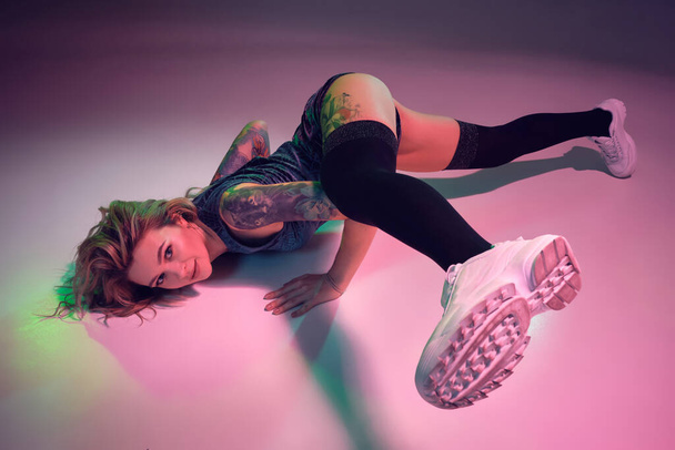 Young beautiful caucasian blonde girl with tattoo on her body, wearing velour blue booty shorts and black stockings dancing twerk lying down with raised leg, pink background - Foto, immagini