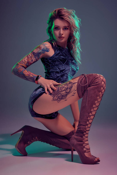 Young beautiful tattooed blonde woman wearing blue velour booty shorts and t-short, maroon suede boots, showing some twerk moves, looks seductive. - Photo, Image