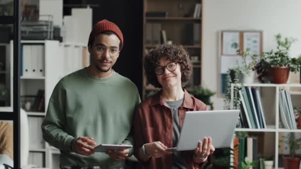Retrato de una joven pareja de desarrolladores de pie con gadgets y sonriendo a la cámara durante su trabajo en equipo en la oficina - Metraje, vídeo