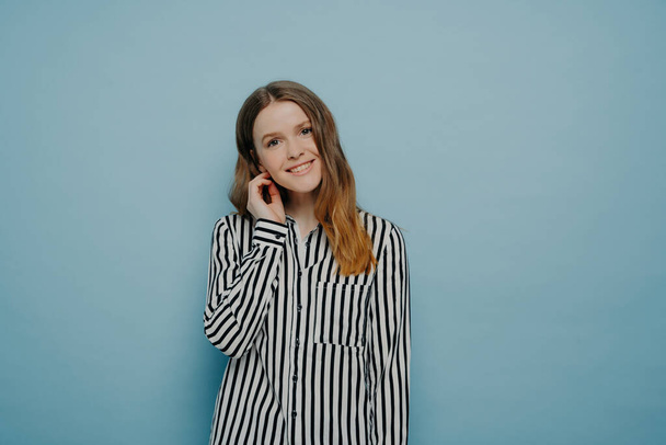 Tender schattig Europees tiener meisje in gestreept wit shirt, kantelen hoofd met haar achter het oor, uiten verlegenheid, poseren in de studio tegen lichtblauwe achtergrond. - Foto, afbeelding