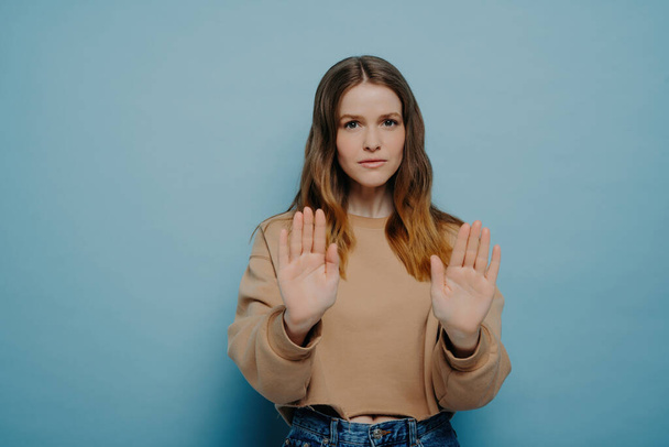 Serious attractive young woman with brown hair in casual wear, holding hands in front, demonstrating a stop gesture while posing on a light blue studio background. - Photo, Image