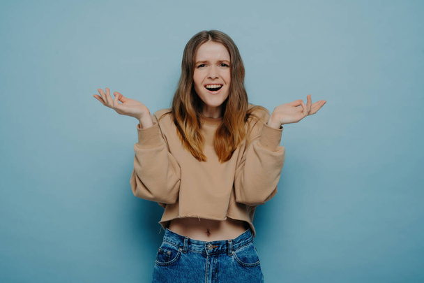 Studio shot of attractive amazed teenage girl in stylish casual outfit, cannot believe her eyes, shrugging shoulders with a surprised face expression, looking at the camera, isolated on a blue background. - Foto, imagen