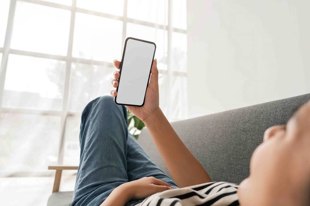 Молодая женщина азиатского отдыха на удобном диване и проведение мобильного телефона с белым экраном. - Фото, изображение